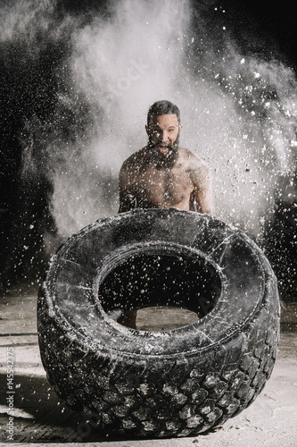 Bearded athlete screaming and pushing big tire in dust on black background photo