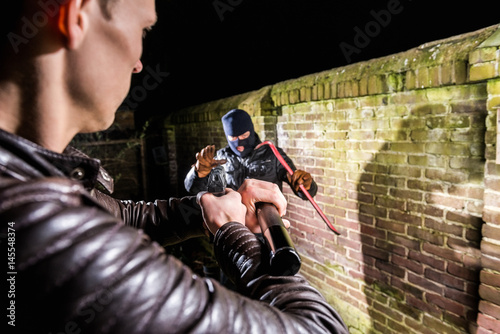 Policeman aiming torch and pistol towards busted scared cracksman by brick wall at night photo