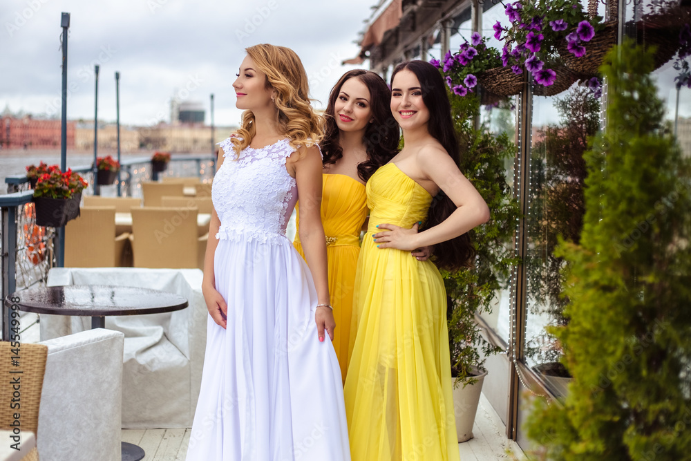 Naklejka premium Beautiful Bride and two bridesmaids in yellow similar dresses together on a summer terrace a sea restaurant