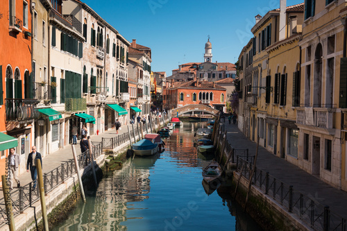 Fototapeta Naklejka Na Ścianę i Meble -  Narrow street with a canal, bridge, boats and tourists in Venice