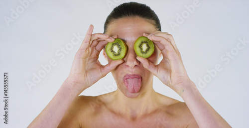 Portrait of a beautiful young girl covers her eyes with a kiwi and shows her smiles, on a white background: Concept: vitamins, fruits, fresh, bio products, summer, spring, spa procedures, skin care.