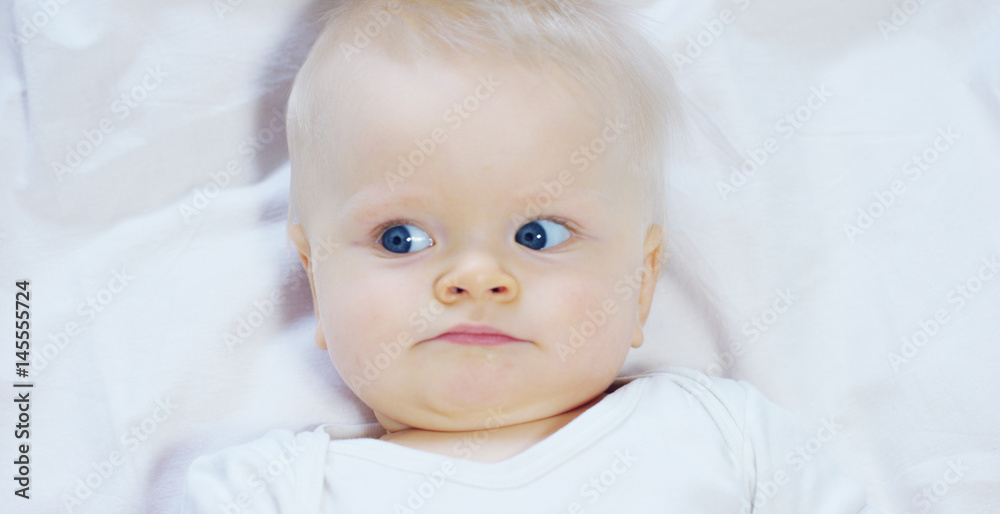 A baby, a boy with large blue eyes and light-colored hair, lies and smiles on a snow-white blanket, looks at her mother, on a white background.