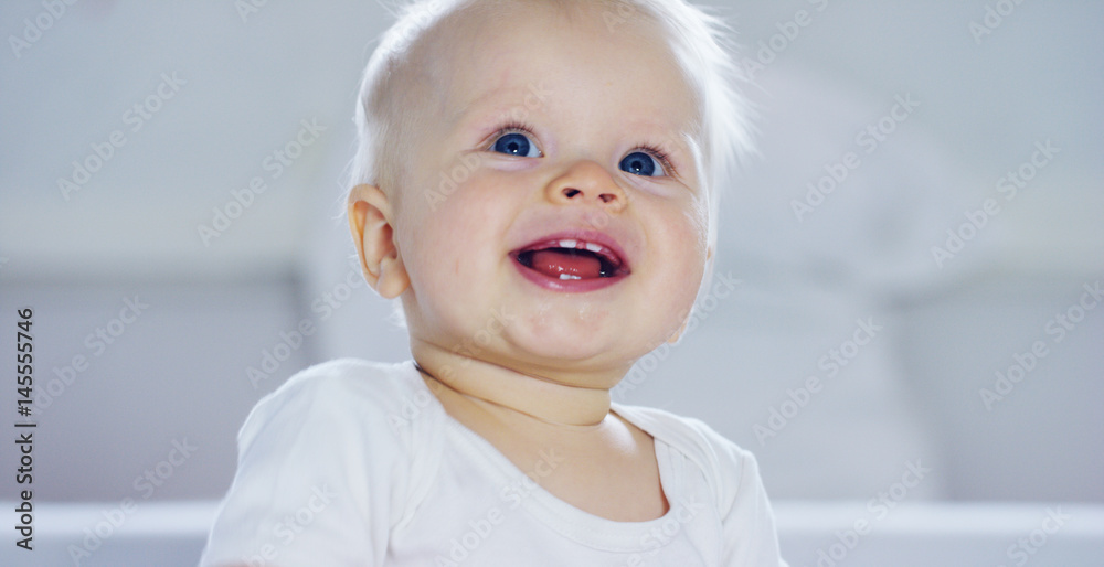 A baby, a boy with large blue eyes and light-colored hair, sits and smiles on a snow-white blanket, looks at her mother, on a white background.