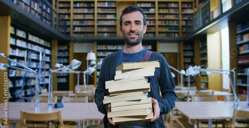 Portrait of a beautiful young man smiling happy in a library holding books after doing a search and after studying. Concept: educational, portrait, library, and studious.