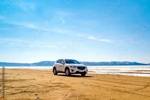 A white car in the spring on a sandy beach in front of a river, mountains, melting ice