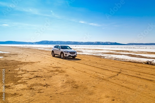 A white car in the spring on a sandy beach in front of a river, mountains, melting ice