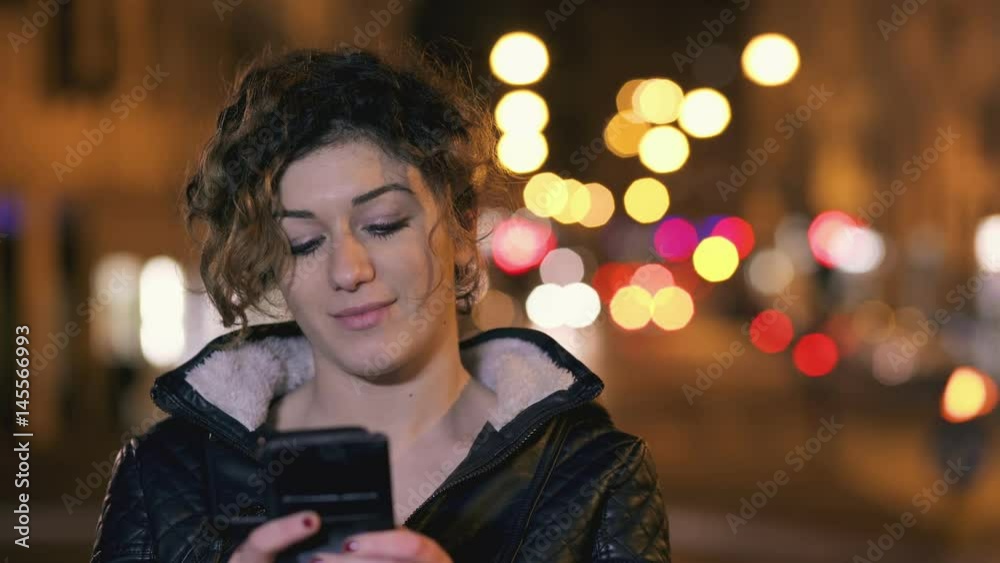 Beautiful girl writes a message with the smartphone, city night in the backgroun Beautiful girl writes a message with the smartphone, city night in the backgroun