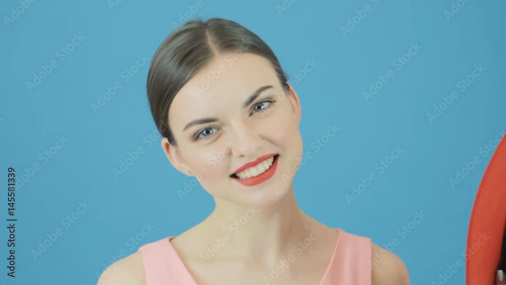 Portrait of Dark-haired Woman Hides Her Face with Red Hat on Blue Background in Studio. Portrait of Dark-haired Woman Hides Her Face with Red Hat on Blue Background in Studio.