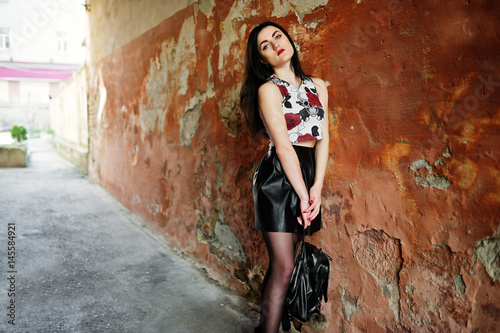 Young goth girl on black leather skirt with backpack posed against grunge wall.