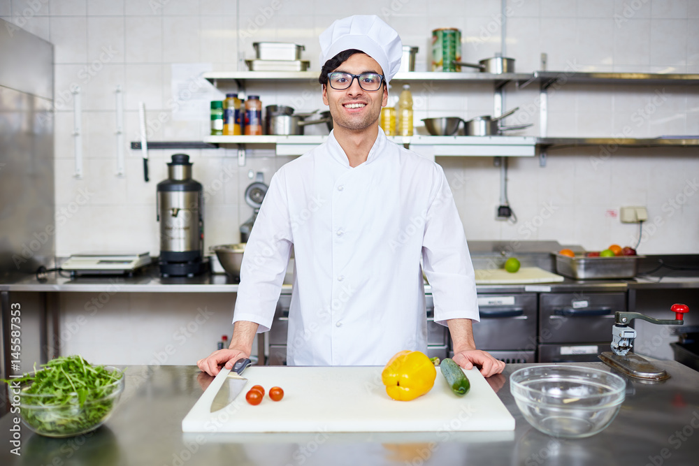Happy young chef in uniform standing by workplace in the kitchen Happy young chef in uniform standing by workplace in the kitchen