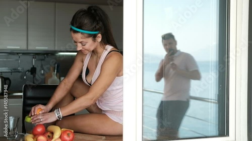 Young woman squeezes orange juice sitting by window in kitchen. Sweet brunette in pink body with bare legs coquettishly settled on table on which stand juicer and fruit. Fascinated by process of