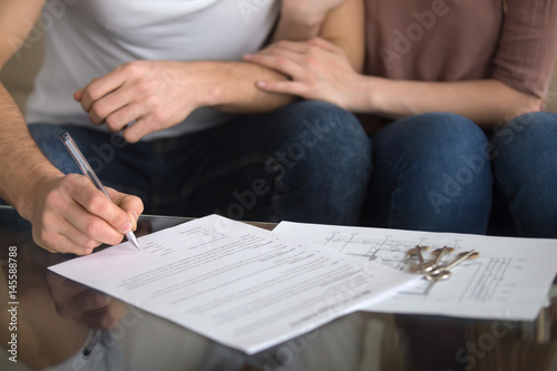 Buying and renting new home concept. Close up of loving couple signing rental agreement or sale purchase contract sitting on couch indoors, starting family life, approved loan to buy real estate