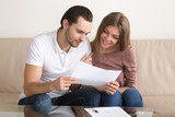 Smiling couple holding papers and carefully studying documents sitting on sofa indoors, reading terms and conditions, reviewing agreement, considering mortgage loan offer, personal insurance