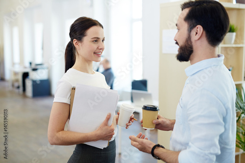 Portrait of smiling young woman talking to colleague during break in office, both holding paper coffee cups
