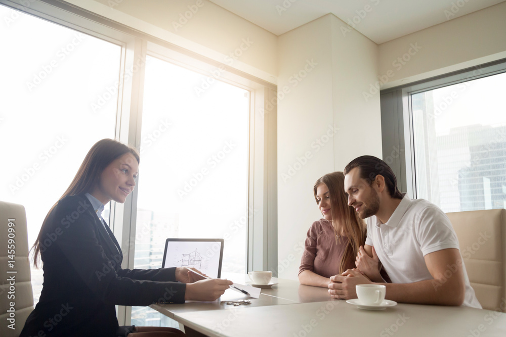 Female realtor meeting with young couple in real estate agency office. Discussing first property investment with broker, construction company manager showing build project, apartment plan on a laptop Female realtor meeting with young couple in real estate agency office. Discussing first property investment with broker, construction company manager showing build project, apartment plan on a laptop