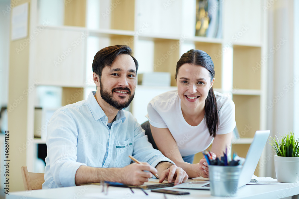 Fototapeta premium Portrait of successful man and woman smiling cheerfully while looking at camera working at desk in office