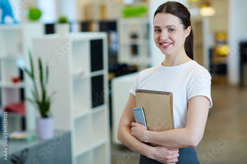 Portrait of young beautiful brunette woman smiling happily while looking at camera standing in modern office photo