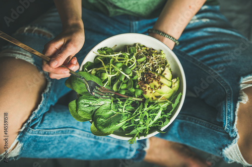 Green vegan breakfast meal in bowl with spinach, arugula, avocado, seeds and sprouts. Girl in jeans holding fork with knees and hands visible, top view. Clean eating, dieting, dieting food concept