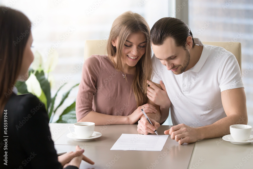 Fototapeta premium Smiling couple agreed to sign prenuptial contract, handsome man putting signature on document while sitting together with his wife, taking bank loan, health insurance, signing financial papers