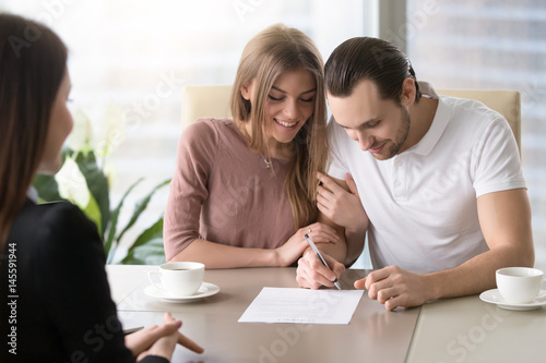 Smiling couple agreed to sign prenuptial contract, handsome man putting signature on document while sitting together with his wife, taking bank loan, health insurance, signing financial papers photo