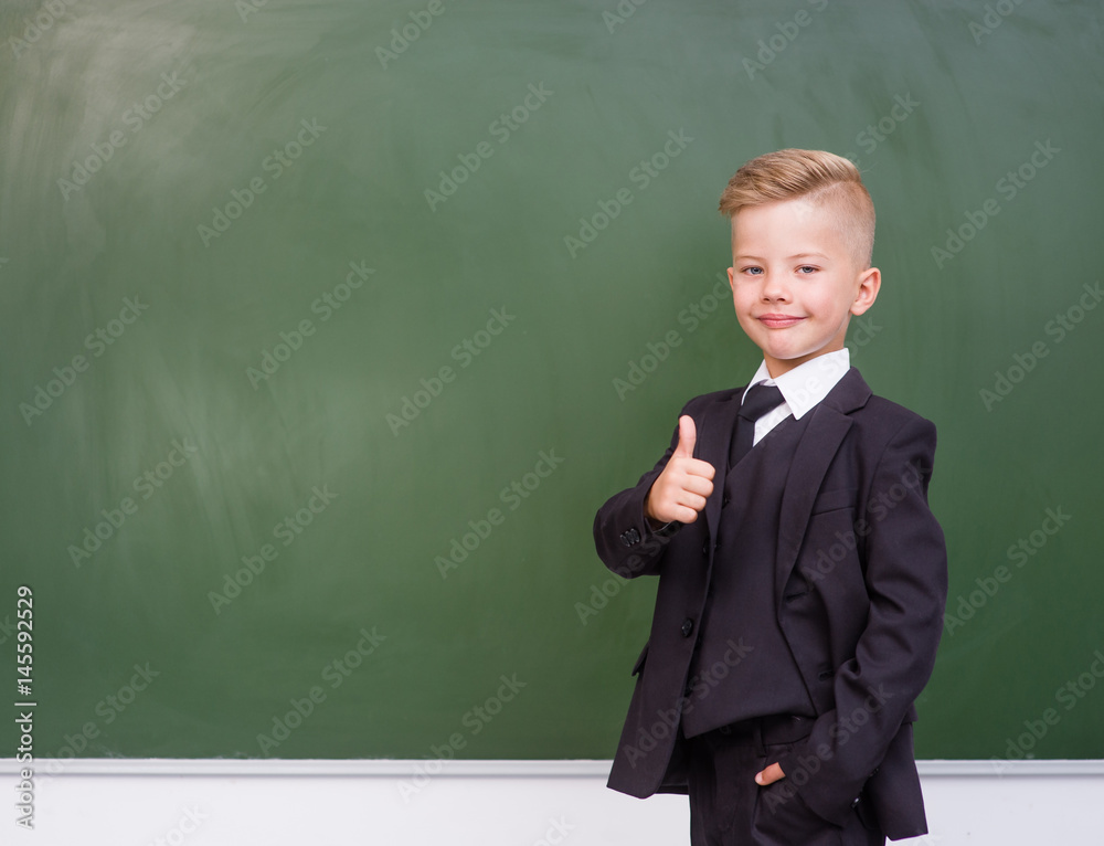 Boy in a business suit showing thumbs up near empty green chalkboard