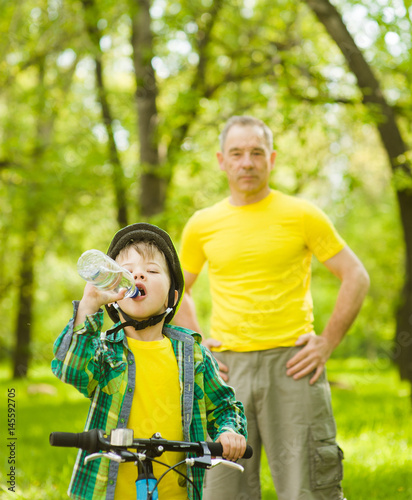 Young boy with a bottle of water is learning to ride a bike with his father