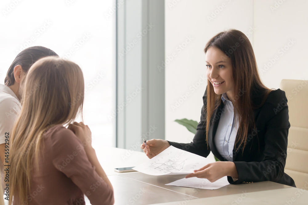 Smiling real estate agent offering to young couple to buy a cottage while meeting at office, presenting technical documentation such as building house plan, telling about advantages of country living Smiling real estate agent offering to young couple to buy a cottage while meeting at office, presenting technical documentation such as building house plan, telling about advantages of country living