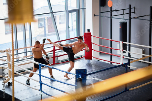 Wide shot of two men fighting in boxing ring: boxer laying high kick hit to head