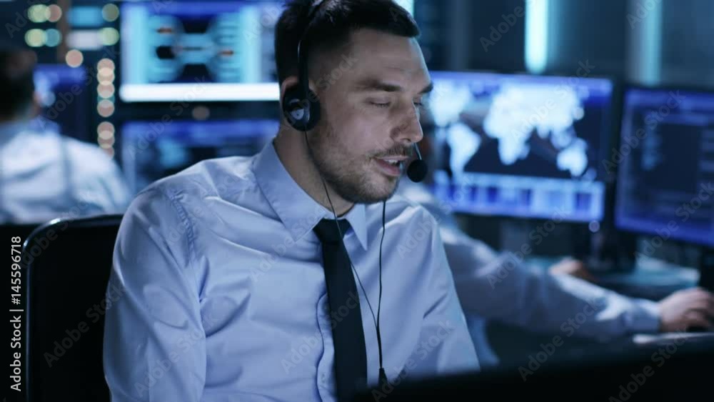 In System Control Center Technical Support Specialist Speaks into Headset. His Colleagues are Working in the Background in a Room Full of Screens. Shot on RED EPIC-W 8K Helium Cinema Camera. In System Control Center Technical Support Specialist Speaks into Headset. His Colleagues are Working in the Background in a Room Full of Screens. Shot on RED EPIC-W 8K Helium Cinema Camera.