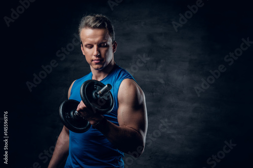 A man dressed in a blue sportswear holds the dumbbell over dark grey background. © Fxquadro