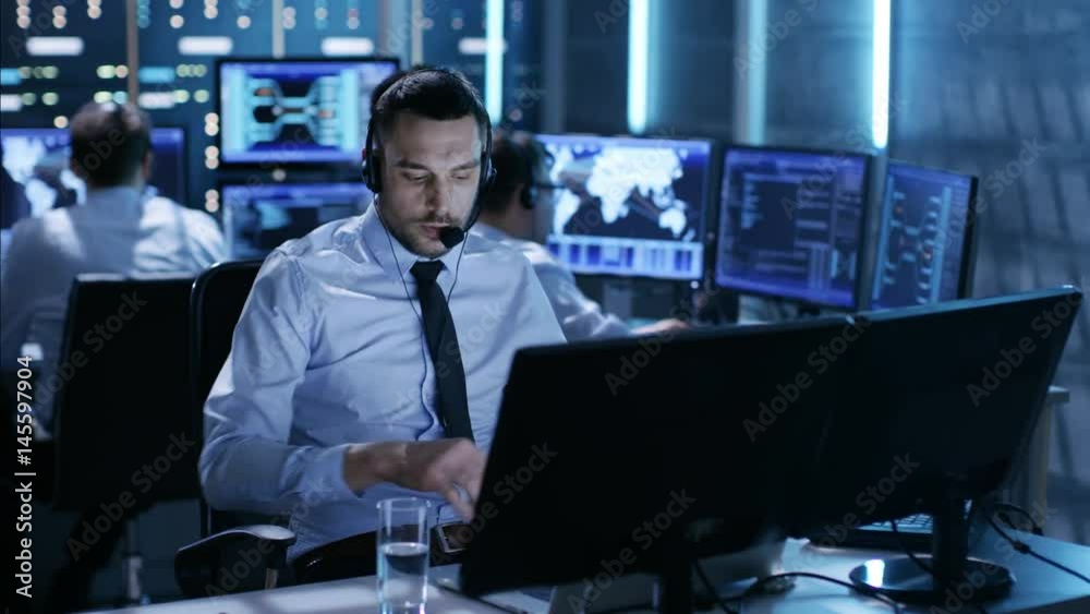 In System Control Center Technical Support Specialist Speaks into Headset while Sitting at His Desk Before Multiple Monitors. His Colleagues are Working in the Background in a Room Full of Displays. In System Control Center Technical Support Specialist Speaks into Headset while Sitting at His Desk Before Multiple Monitors. His Colleagues are Working in the Background in a Room Full of Displays.
