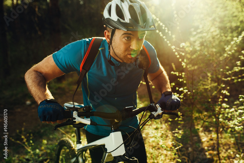 Attractive serious male biker wearing blue sports clothing, helmet and gloves, having confident and self-determined look on forest sunset or sunrise. Travel, sport, extreme concept.