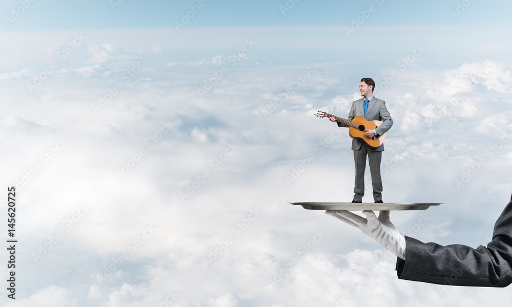 Businessman on metal tray playing acoustic guitar against blue sky background Businessman on metal tray playing acoustic guitar against blue sky background