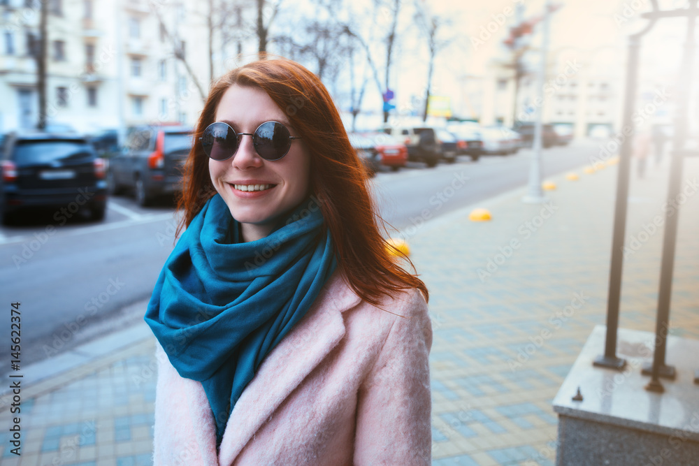 Fototapeta premium Red haired beautiful girl is walking by the street in a pink coat and blue scarf, with sunglasses.