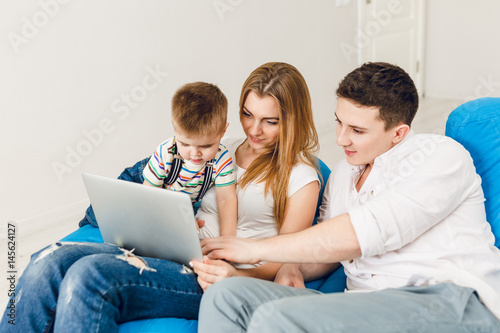 Young family of two parents and one boy child sit on blue sack chairs. They wear white t-shirt and jeans. Mom holds a laptop and a boy plays on it. They sit in a studio with white walls.