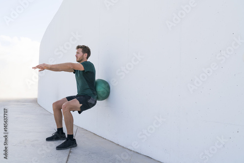 Strength training man doing squats using medicine ball rolling on wall squatting at fitness centre. Workout squat bodyweight exercises using medicine ball. Fitness athlete working out.