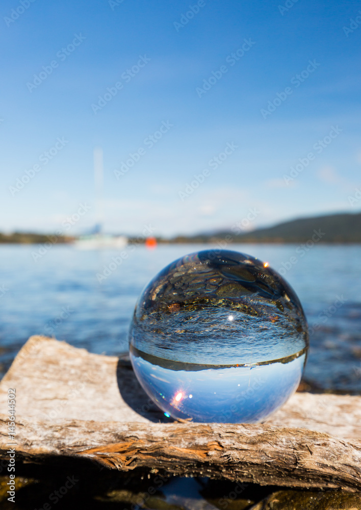 Glaskugel auf Baumstamm am See mit Segelboot im Hintergrund