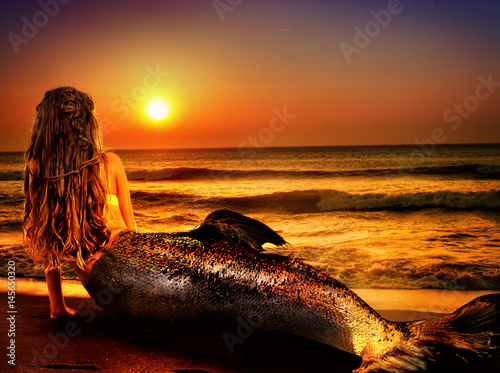 Water nymph mermaid woman dreams on sea sandy beach. Fairy nixie girl looks at tropical setting sun. Fantasy sunset painted undine, sky and sea waves in gold. Magical fish tail of naiad shining squama