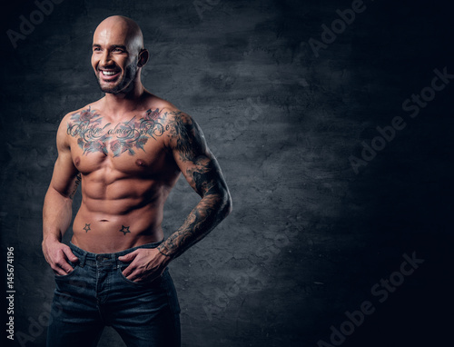 Shaved head, muscular male with tattoos on his torso over grey vignette background.