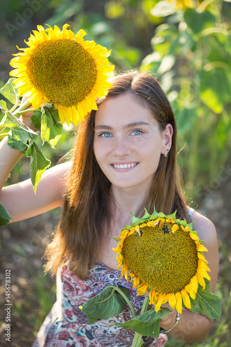 Cute beautiful young girl lady woman sitting on a field with big sunflowers.Brunette with blue eyes wearing colorful dress italian leghorn sun bonnet hat on dale lea of green yellow blooms blossom. photo