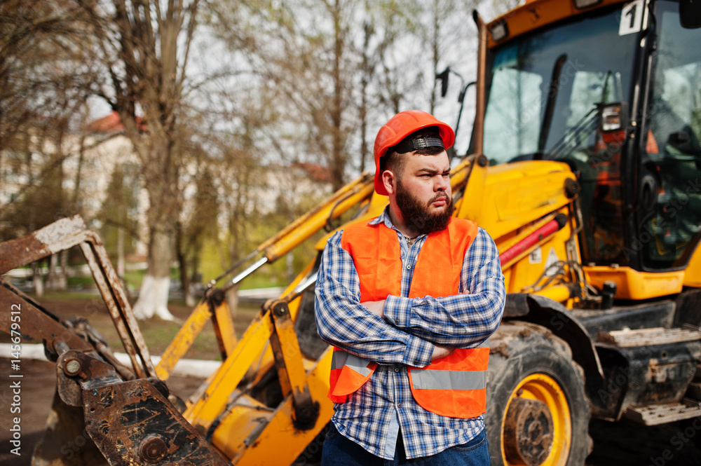 Brutal beard worker man suit construction worker in safety orange helmet against traktor.