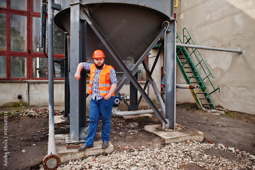 Brutal beard worker man suit construction worker in safety orange helmet stay near big industrial barrel . Brutal beard worker man suit construction worker in safety orange helmet stay near big industrial barrel .