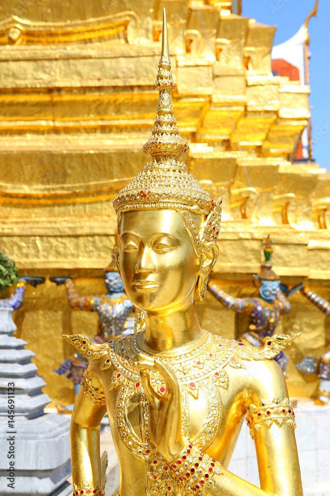 Fototapeta premium Golden Kinnaree holding flower against pagoda background on display at wat pra kaew in Bangkok, Thailand.