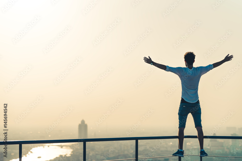 Fototapeta premium Freedom or success concept. White man standing at the edge of rooftop, stretching arms. Sunset on cityscape background