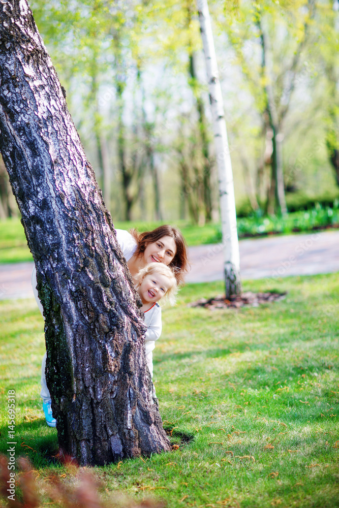 Mother and daughter playing hide and seek in the spring forest. They look out from behind the birch and smile cheerfully Mother and daughter playing hide and seek in the spring forest. They look out from behind the birch and smile cheerfully