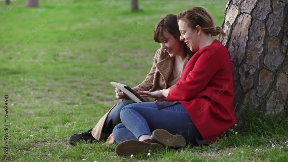 Two relaxed friends sitting under a tree use the tablet Two relaxed friends sitting under a tree use the tablet
