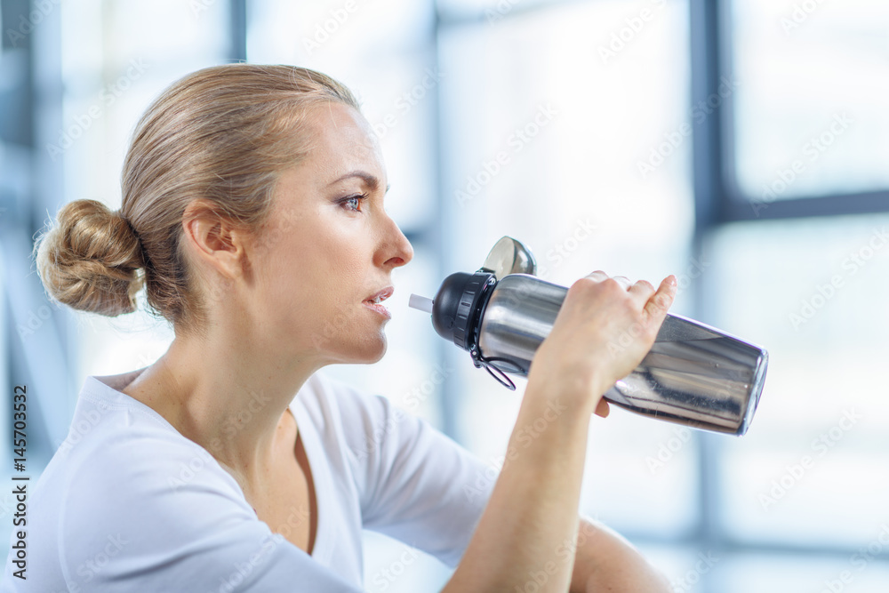 tired sportswoman drinking water from sport bottle in sports center tired sportswoman drinking water from sport bottle in sports center