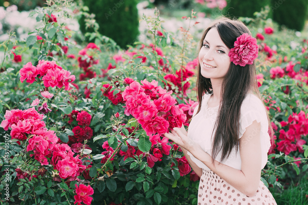 Красивая девушка с длинными волосами в платье стоит возле розовых роз в саду 