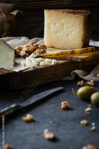 Cheese platter of chopped Spanish hard cheese manchego and sliced Italian pecorino toscano) on wooden board, with green olives in glass jar and walnuts on dark rustic background photo