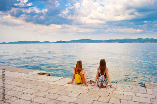 Two young girls with backpacks sitting on the stone near the sea shore, their hands leaning against stone pavement, looking at the hill range in the distance. Town of Zadar, Croatia